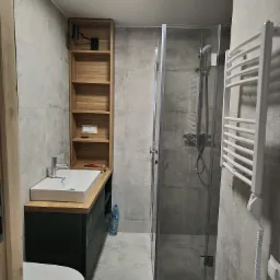 Nowoczesna łazienka w szarościach z kabiną prysznicową, umywalką nablatową na szafce w kolorze grafitowym, drewnianym regałem i grzejnikiem drabinkowym.