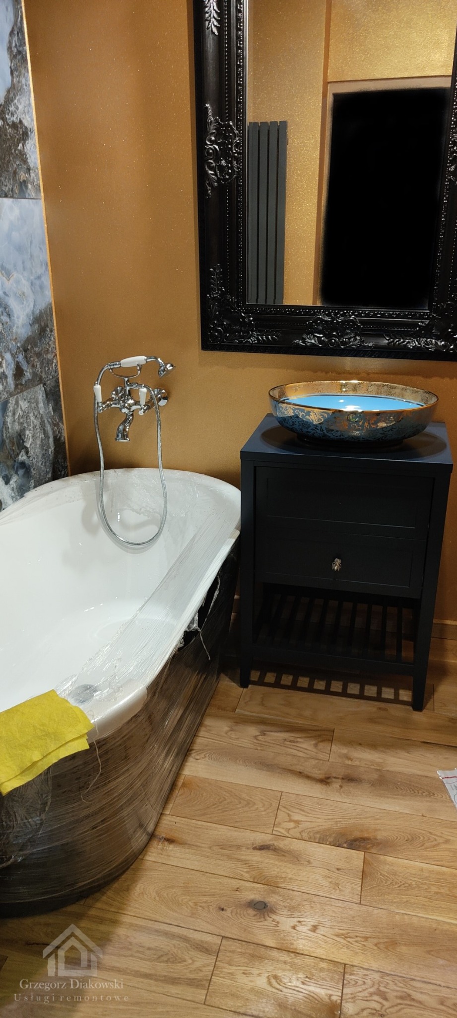 Biała wanna wolnostojąca w folii ochronnej z żółtą ściereczką, umywalka w stylu retro z niebieską wodą na czarnej szafce, lustro w ozdobnej ramie, złota ściana i drewniana podłoga podczas prac...