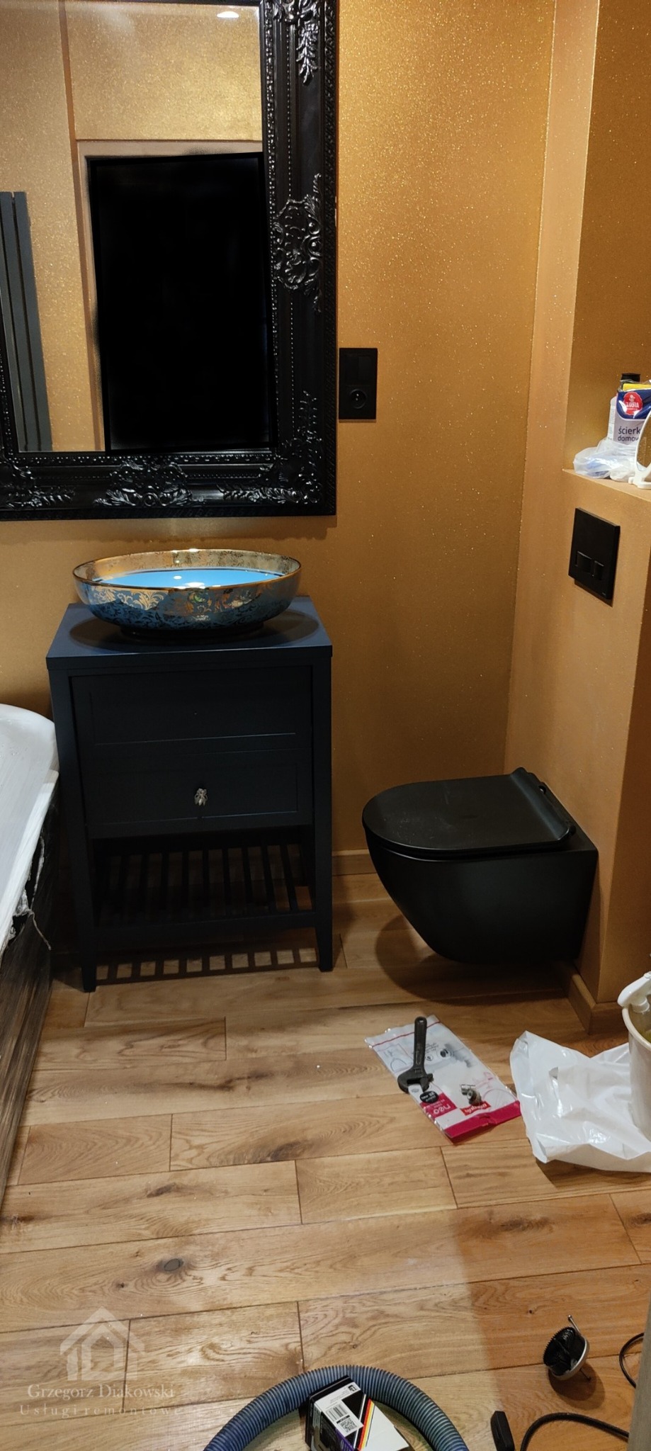 Nowoczesna łazienka z brokatową ścianą, wiszącą toaletą w kolorze czarnym, umywalką w kolorze niebieskim z elementami złota na szafce również w kolorze czarnym oraz czarnym lustrem w ozdobnej ramie...