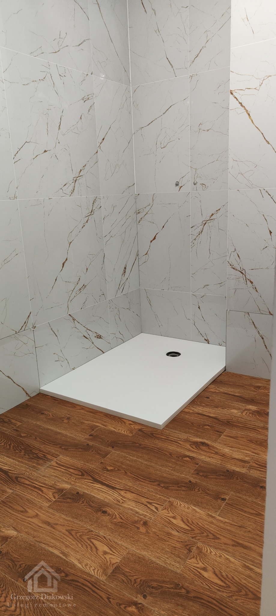 Wykończony brodzik z odpływem w rogu łazienki, obłożony białymi płytkami imitującymi marmur z brązowymi żyłami, na podłodze panele imitujące drewno.