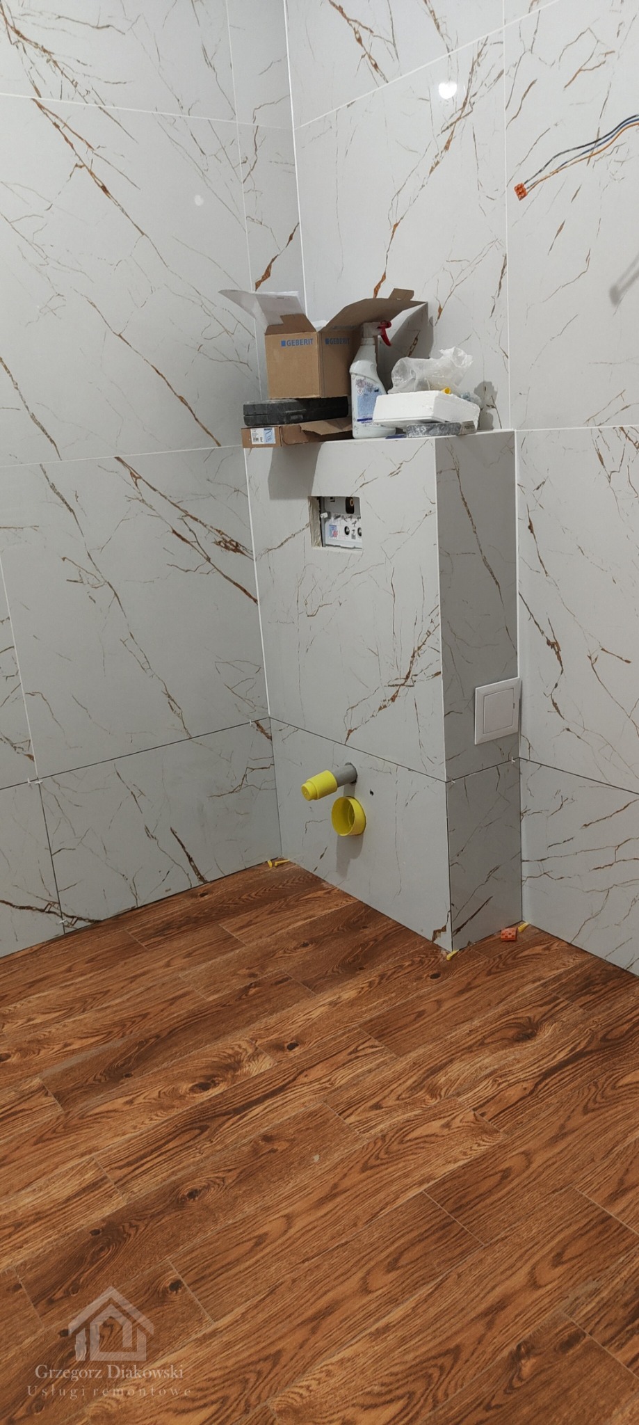 Łazienka w trakcie prac wykończeniowych: ułożone płytki imitujące marmur na ścianach, drewnopodobna podłoga, widoczne żółte rury kanalizacyjne i pudełka po armaturze.