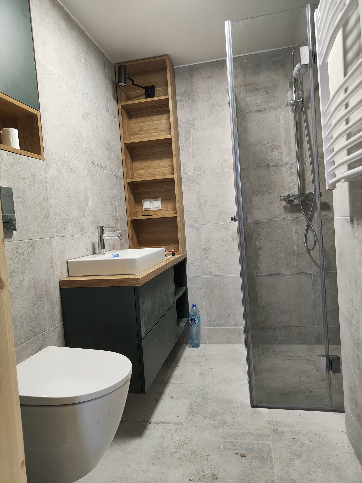 Nowoczesna łazienka z szarą płytką, umywalką nablatową na ciemnozielonej szafce, drewnianym regałem z oświetleniem, kabiną prysznicową i podwieszaną toaletą.