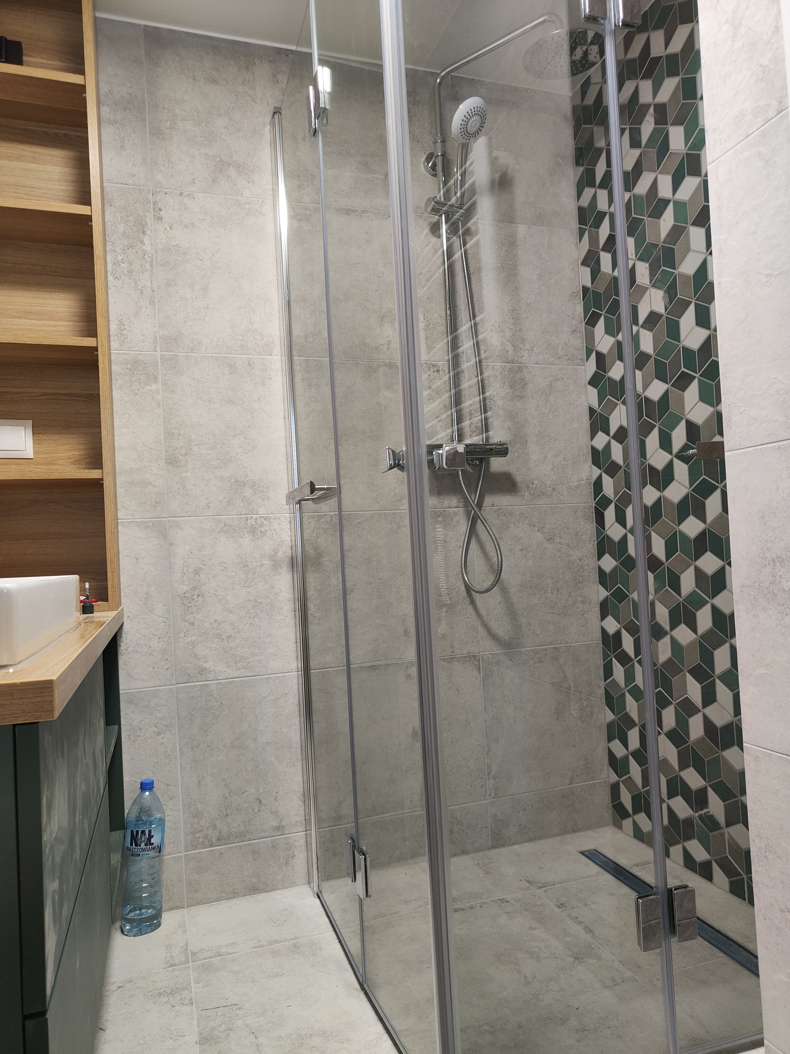 Nowoczesna kabina prysznicowa z geometryczną mozaiką na ścianie, szklane drzwi z chromowanymi zawiasami, szara płytka podłogowa i butelka wody mineralnej przy umywalce.