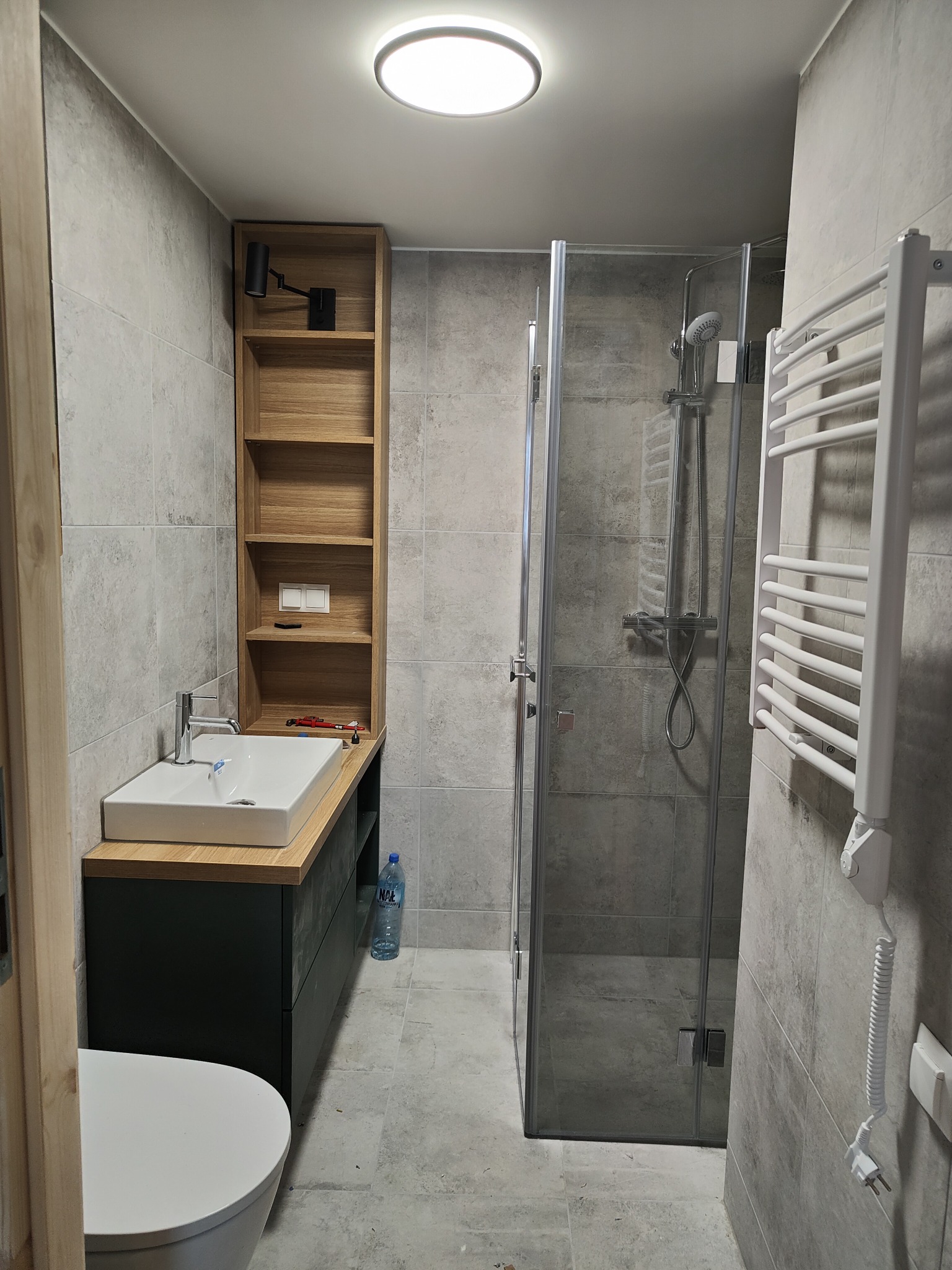 Nowoczesna łazienka w szarościach z kabiną prysznicową, umywalką nablatową na szafce w kolorze grafitowym, drewnianym regałem i grzejnikiem drabinkowym.