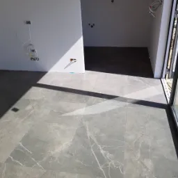 Ułożona szara podłoga z płytek imitujących marmur, widoczne przygotowane instalacje elektryczne w pomieszczeniu w trakcie remontu, cień rzucany przez okno.