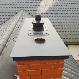 Kompletny komin z cegły z czarnym daszkiem i wkładem kominowym na tle szarej dachówki, widoczne narzędzia i rękawice robocze.