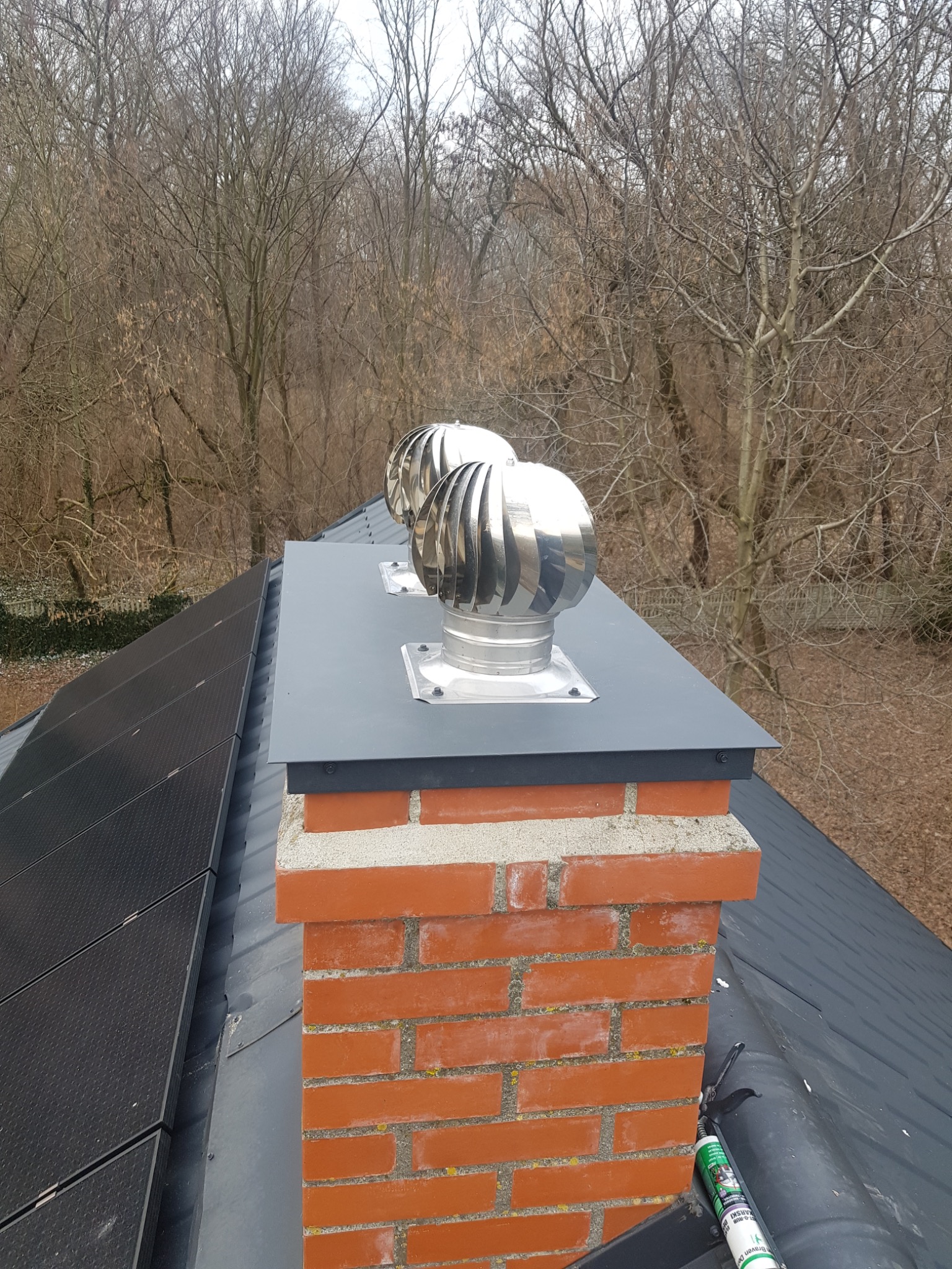 Ceglany komin z dwoma metalowymi nasadami obrotowymi na dachu z panelami słonecznymi w tle drzew bez liści