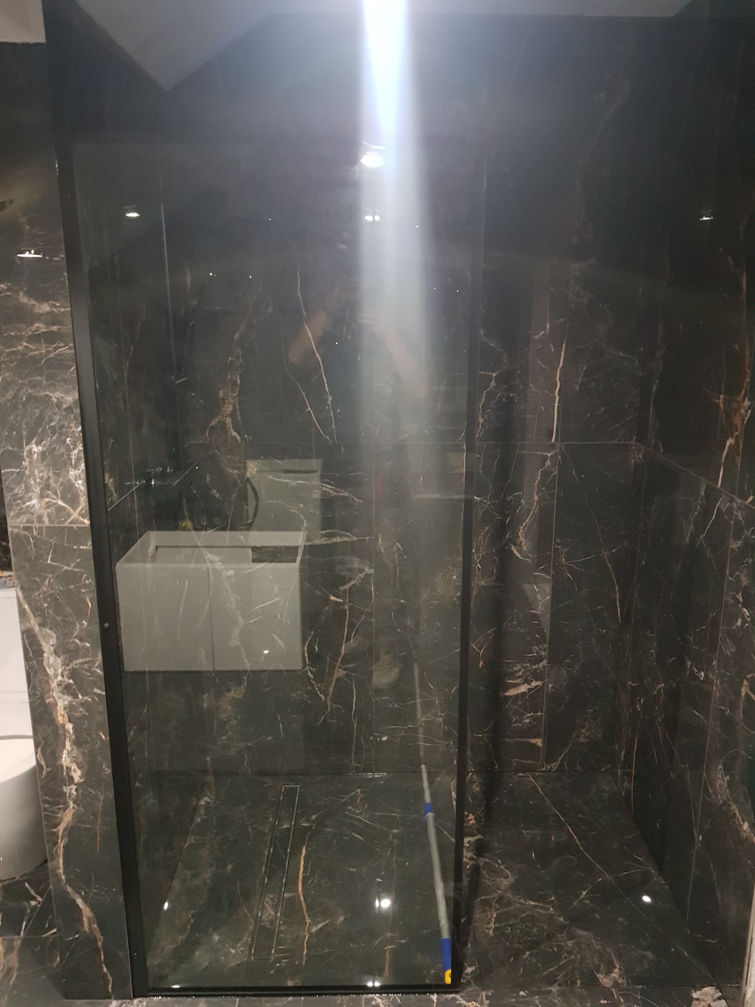 Nowoczesna kabina prysznicowa z czarnymi profilami i marmurowymi płytkami w ciemnych odcieniach, widoczny odpływ liniowy w podłodze i umywalka.