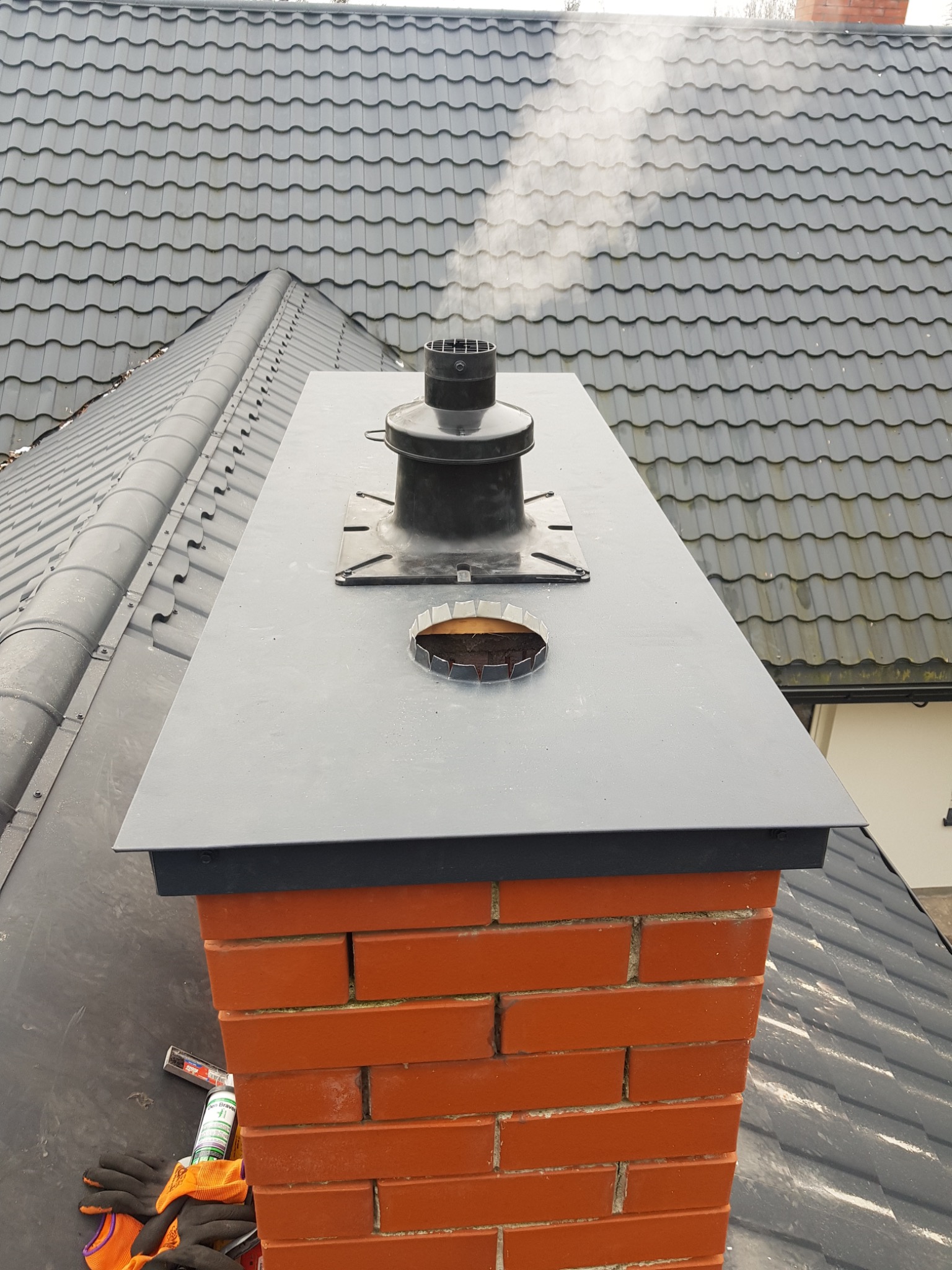 Kompletny komin z cegły z czarnym daszkiem i wkładem kominowym na tle szarej dachówki, widoczne narzędzia i rękawice robocze.
