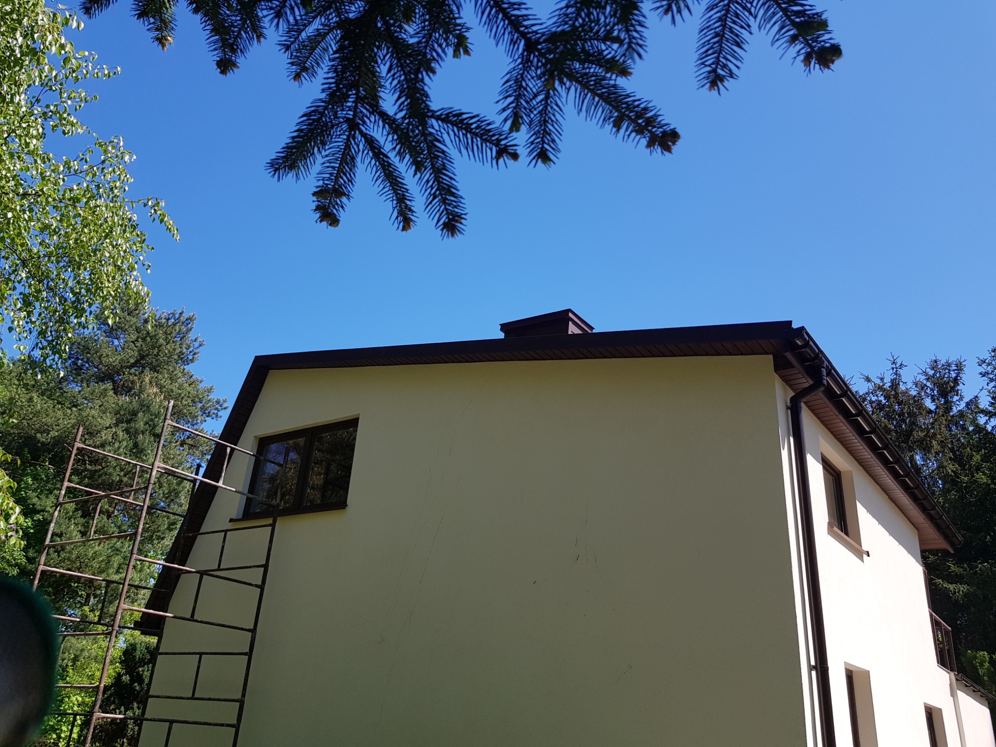 Elewacja domu jednorodzinnego w trakcie prac, widoczne rusztowanie, fragmenty zieleni w tle i błękitne niebo