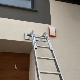 Drabina oparta o ścianę domu podczas montażu zewnętrznej sygnalizacji alarmowej, widoczna biało-czerwona obudowa sygnalizatora.
