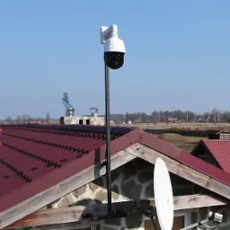 Kamera przemysłowa PTZ na wysokim maszcie, zamontowana na dachu domu z czerwoną dachówką, widok na wieś w tle.