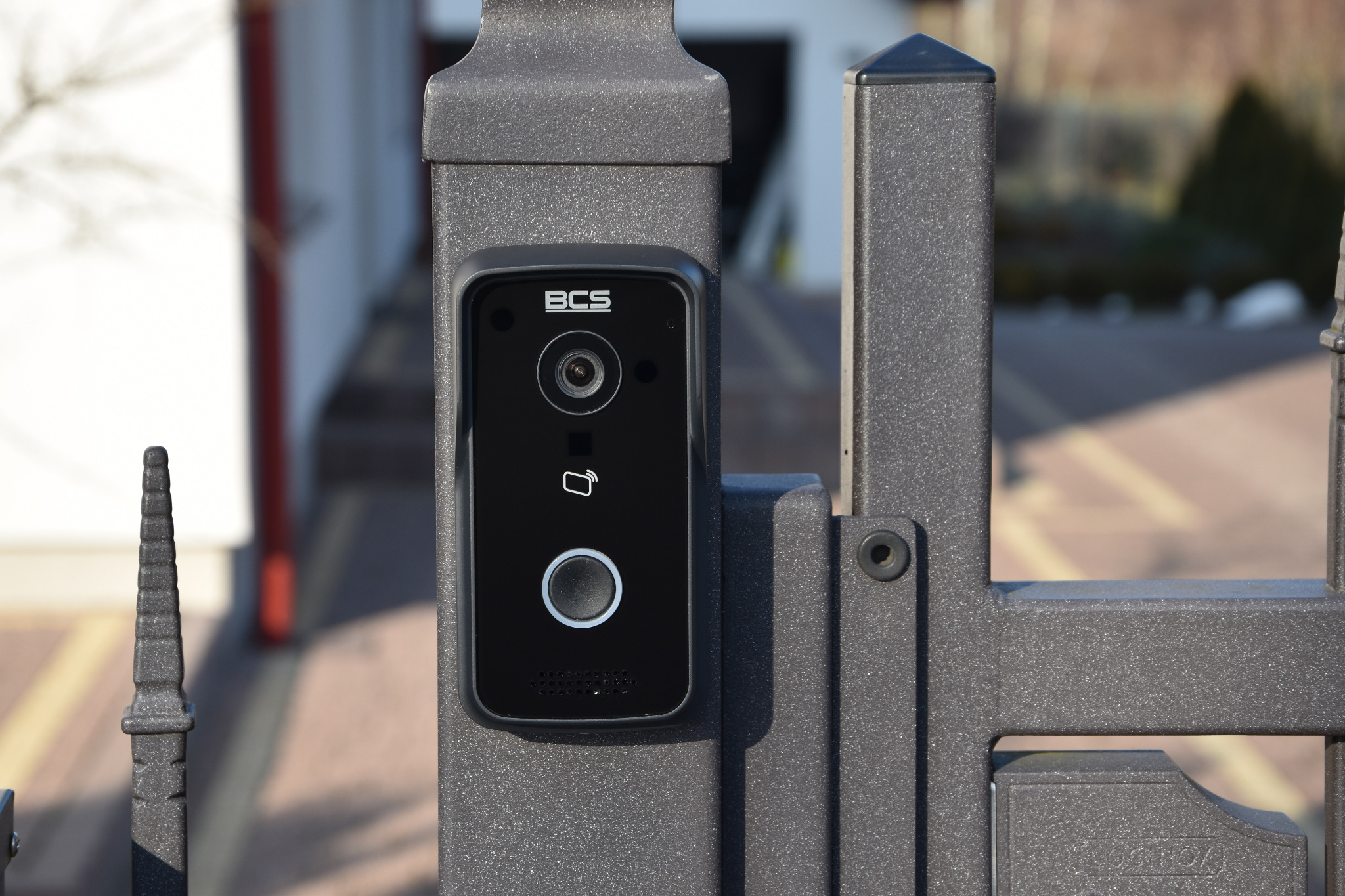 Czarny panel wideodomofonu BCS zamontowany na szarej, metalowej bramie wjazdowej, widok z bliska na urządzenie z widocznym obiektywem kamery i przyciskiem dzwonka.