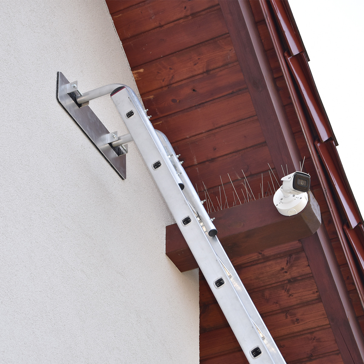 Montaż kamery monitoringu na drewnianej belce pod dachem, widoczna drabina oparta o ścianę i kolce przeciw ptakom.