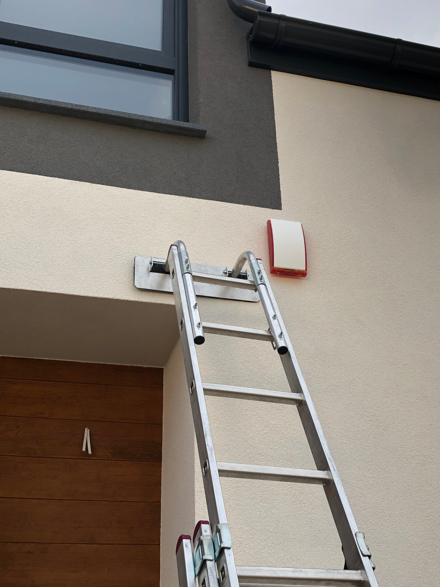 Drabina oparta o ścianę domu podczas montażu zewnętrznej sygnalizacji alarmowej, widoczna biało-czerwona obudowa sygnalizatora.