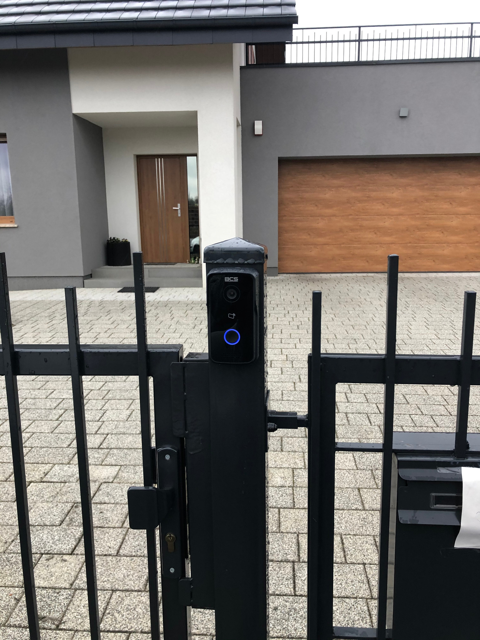 Czarny wideodomofon BCS zamontowany na metalowej bramie posesji z nowoczesnym domem w tle, widoczny podświetlany przycisk i czytnik kart.