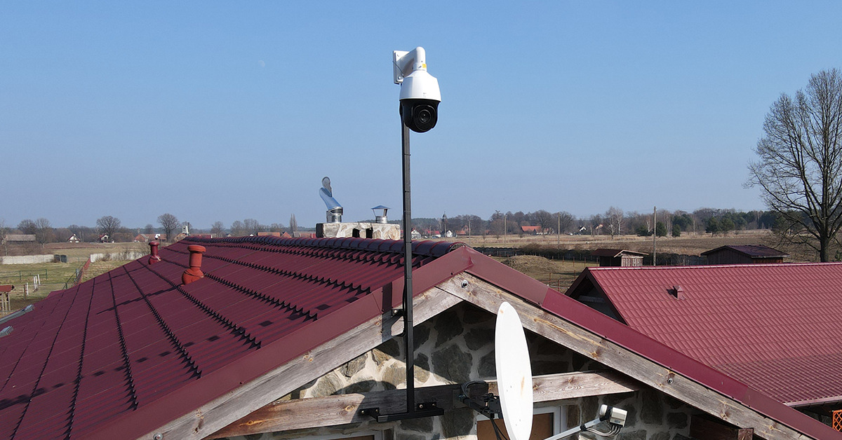 Kamera przemysłowa PTZ na wysokim maszcie, zamontowana na dachu domu z czerwoną dachówką, widok na wieś w tle.