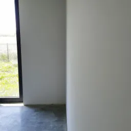 Surowe wnętrze: gładkie, białe ściany, szary betonowy podkład, czarna rama okienna z widokiem na zieleń na zewnątrz, otwór pod oświetlenie w suficie.