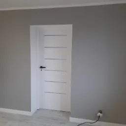 Świeżo pomalowane na szaro ściany z białą, nowoczesną stolarką drzwiową z poziomymi frezowaniami, widoczne gniazdka elektryczne w trakcie montażu.