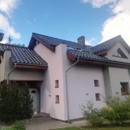 Dwupiętrowy dom po odświeżeniu elewacji w odcieniach szarości i pudrowego różu, z ciemną dachówką i widocznym fragmentem trawnika.