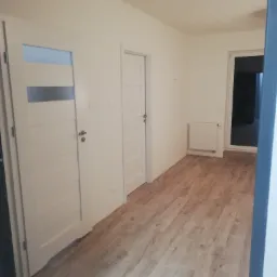 Wnętrze nowo wykończonego pokoju z panelami podłogowymi w odcieniach brązu, białymi ścianami i dwiema białymi, wewnętrznymi drzwiami z przeszkleniami oraz grzejnikiem.