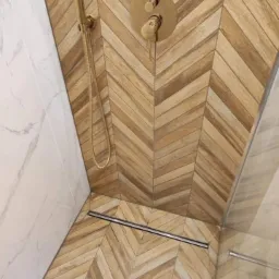 Nowoczesna kabina prysznicowa z drewnopodobnymi płytkami ułożonymi w jodełkę na ścianach i podłodze, złota armatura prysznicowa i odpływ liniowy.