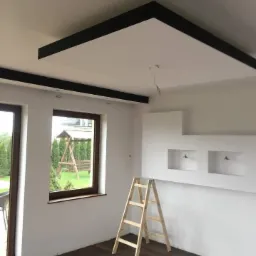 Wnętrze pokoju z drewnianą podłogą, białymi ścianami, oknem z widokiem na ogród, drewnianą drabiną i podwieszanym sufitem z czarnymi elementami dekoracyjnymi.