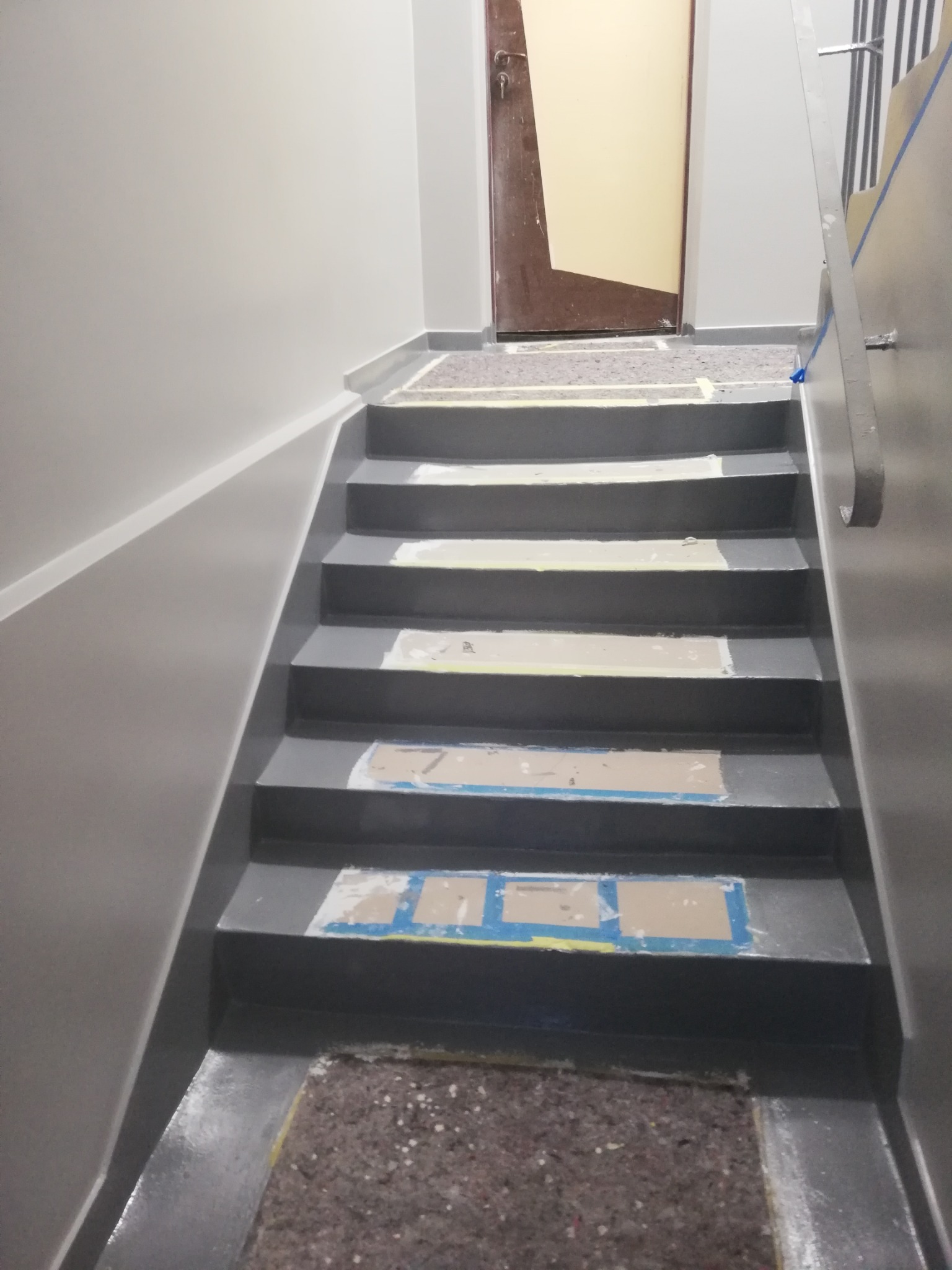 Klatka schodowa w trakcie odświeżania, zabezpieczona taśmą malarską i tekturą, świeżo pomalowane ściany na szaro.
