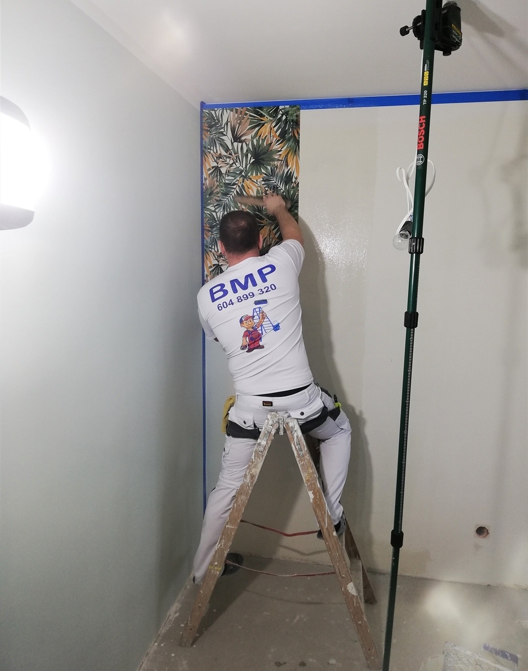 Pracownik na drabinie wygładza wzorzystą tapetę na ścianie, zabezpieczonej niebieską taśmą malarską, widoczny laserowy niwelator Bosch na statywie.