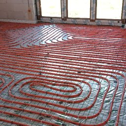 HYDRO-MONTER USLUGI HYDRAULICZNE - Pomarańczowe rury ogrzewania podłogowego ułożone w pętle na siatce z folią odblaskową w surowym wnętrzu z oknami.