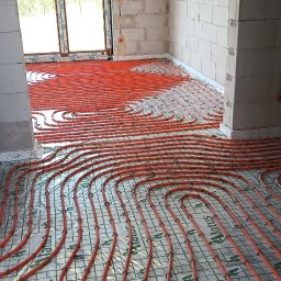 HYDRO-MONTER USLUGI HYDRAULICZNE - Pomarańczowe rury ogrzewania podłogowego zamocowane na folii izolacyjnej z nadrukiem Alatus, widok na surowy stan ścian z betonu komórkowego i okno.