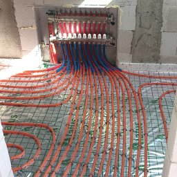 HYDRO-MONTER USLUGI HYDRAULICZNE - Rozdzielacz centralnego ogrzewania z rurami w kolorze czerwonym i niebieskim, ułożonymi na podłodze z siatką i folią izolacyjną, przygotowanymi do zalania betonem.