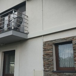 HYDRO-MONTER USLUGI HYDRAULICZNE - Zewnętrzna jednostka klimatyzacji zamontowana na ścianie domu z balkonem i oknem obłożonym kamieniem dekoracyjnym.