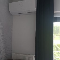 HYDRO-MONTER USLUGI HYDRAULICZNE - Biały klimatyzator Bosch zamontowany na ścianie w pokoju, widoczny grzejnik, zasłony i zabawki na kanapie.