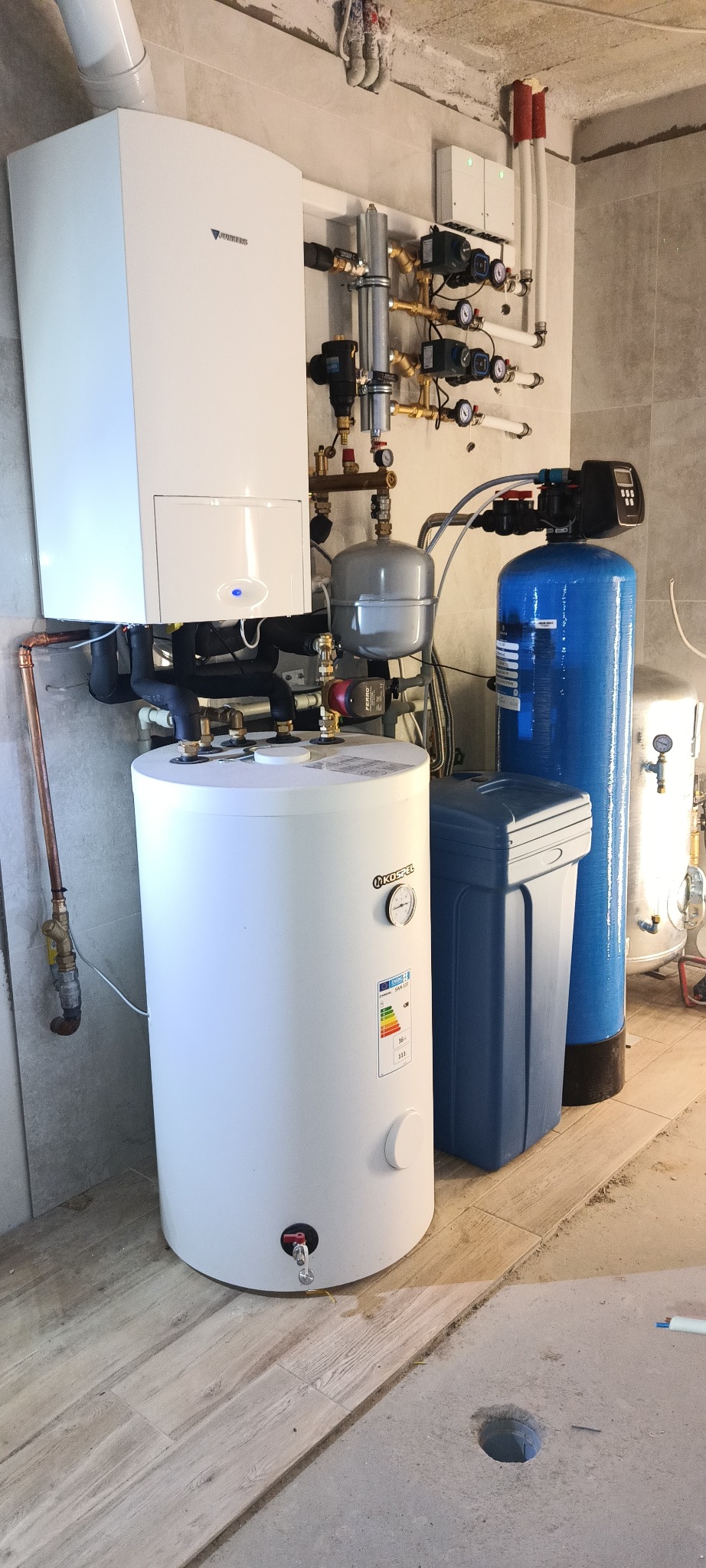 Kotłownia z białym piecem gazowym Junkers, zbiornikiem Kospel, niebieskim filtrem wody i rozbudowaną instalacją hydrauliczną z licznymi zaworami i manometrami, na tle szarych płytek.