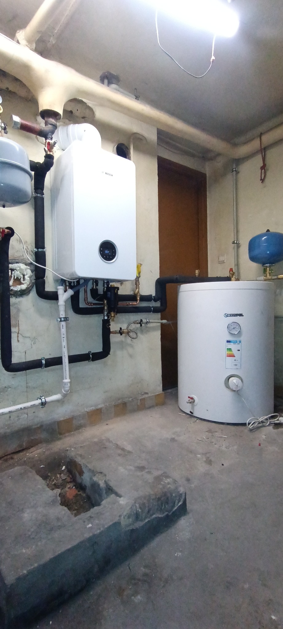 Nowy, biały piec gazowy Bosch zamontowany na ścianie w piwnicy, z podłączonymi rurami miedzianymi i czarnymi, izolowanymi rurami. Obok biały zasobnik ciepłej wody Kospel, widoczne rury instalacji...