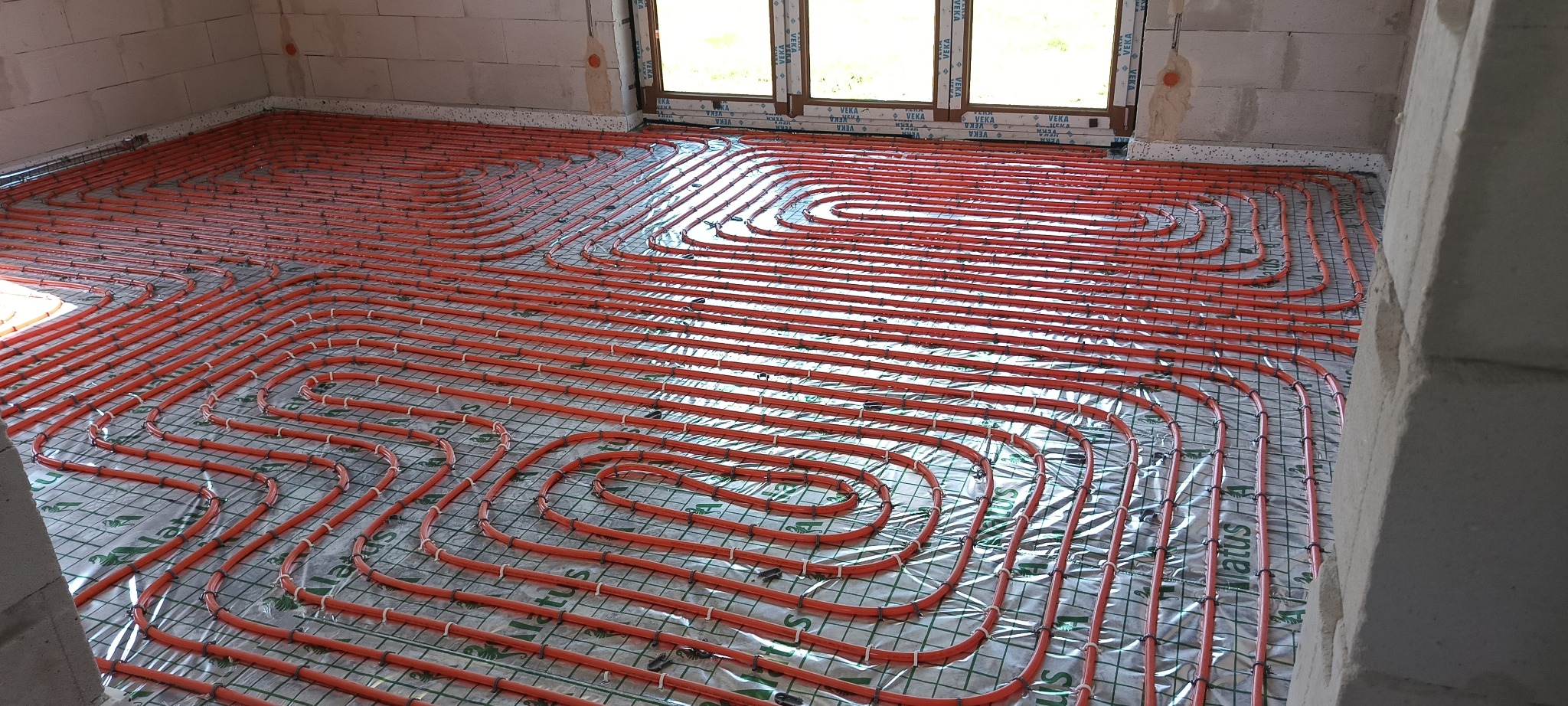 Pomarańczowe rury ogrzewania podłogowego ułożone w pętle na siatce z folią odblaskową w surowym wnętrzu z oknami.