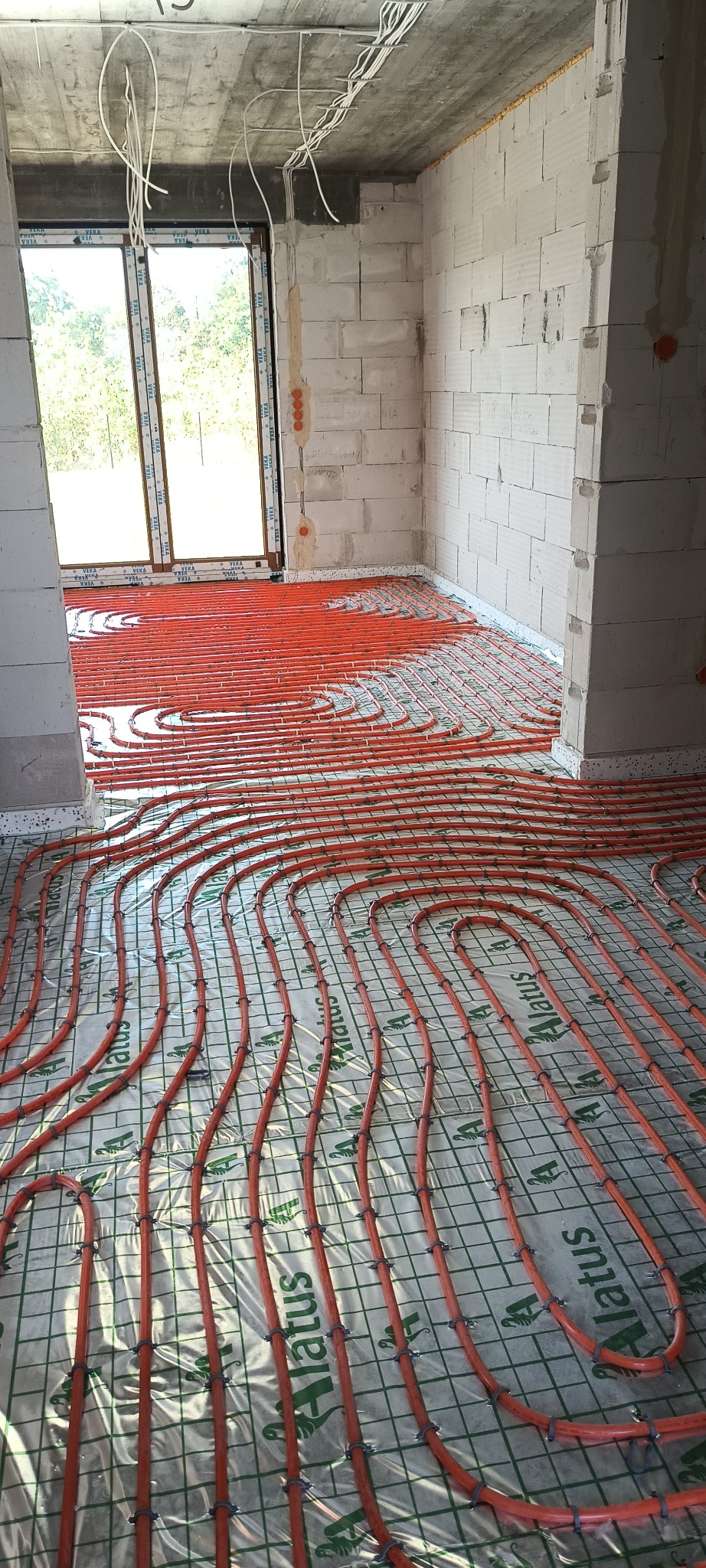 Pomarańczowe rury ogrzewania podłogowego zamocowane na folii izolacyjnej z nadrukiem Alatus, widok na surowy stan ścian z betonu komórkowego i okno.