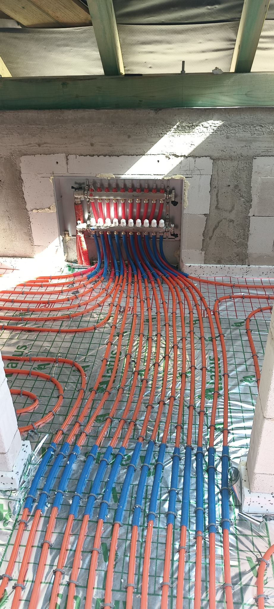 Rozdzielacz centralnego ogrzewania z rurami w kolorze czerwonym i niebieskim, ułożonymi na podłodze z siatką i folią izolacyjną, przygotowanymi do zalania betonem.