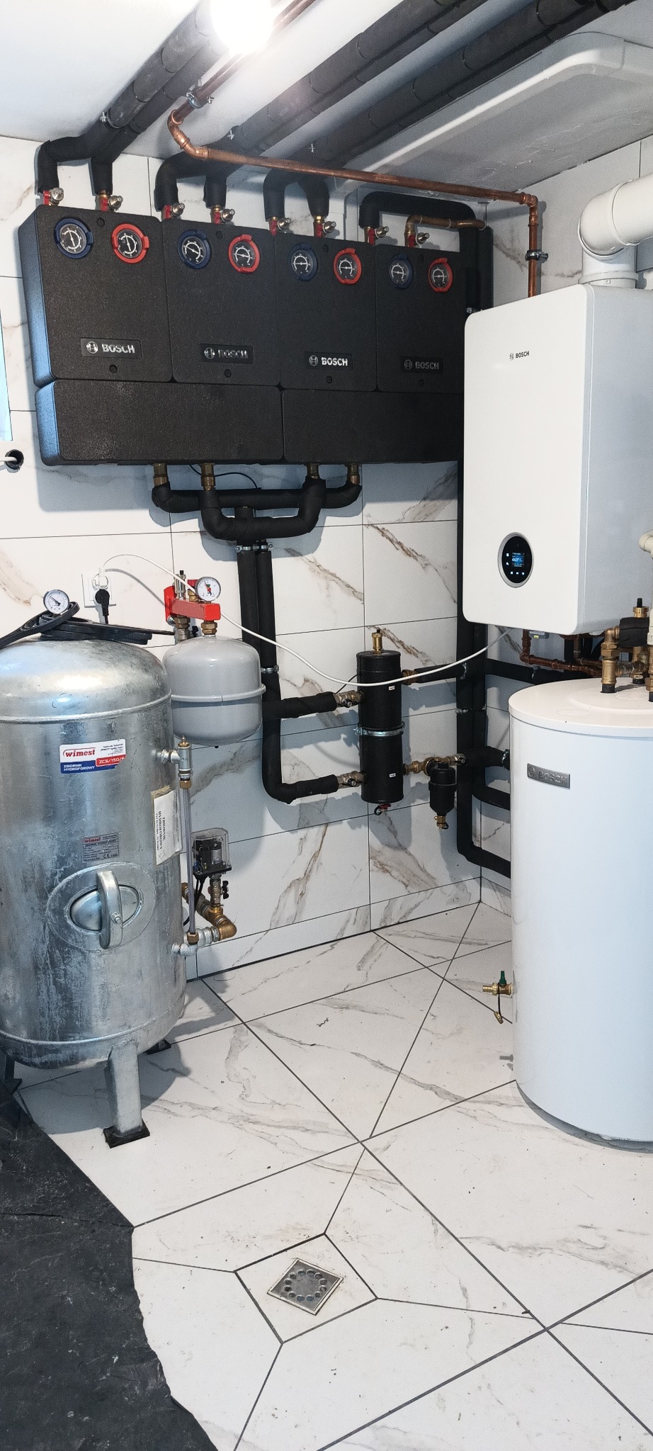 Kompleksowa kotłownia z szeregiem czarnych modułów grzewczych Bosch, białym piecem gazowym, srebrnym zbiornikiem hydroforowym Wimest i czarnymi rurami instalacji na tle białych marmurkowych płytek...