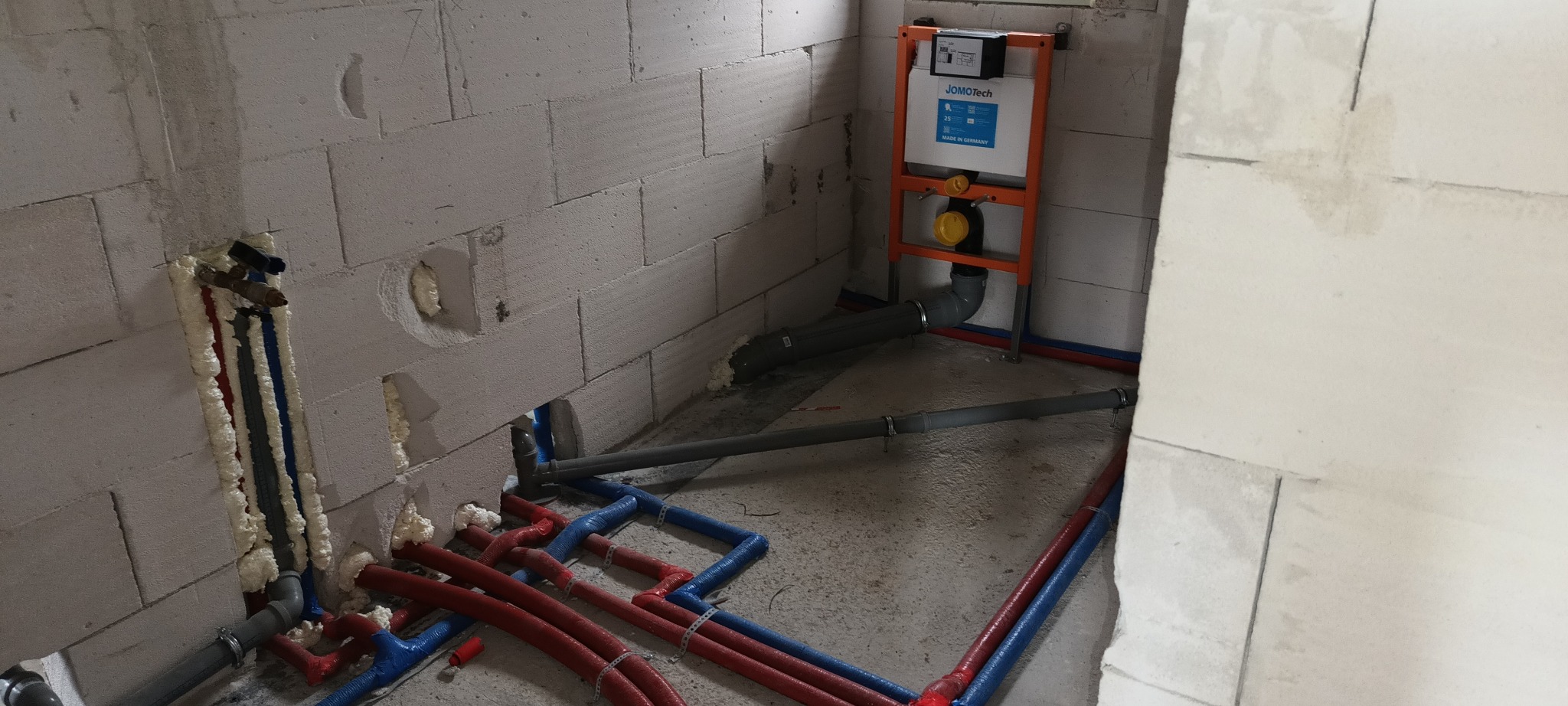 Instalacja podtynkowa WC z widocznymi rurami doprowadzającymi wodę, zabezpieczonymi pianką montażową w surowym pomieszczeniu z bloków betonowych.