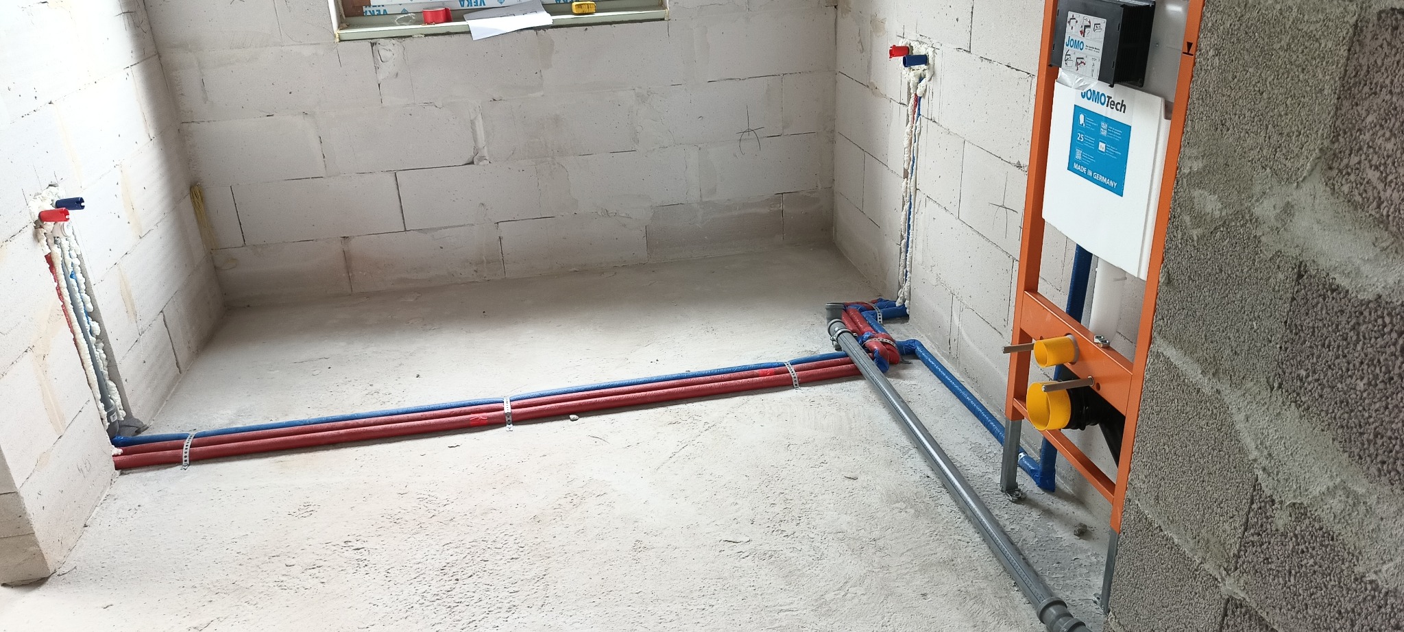 Surowy stan łazienki w trakcie prac instalacyjnych: widoczne rury doprowadzające wodę, stelaż podtynkowy WC JOMO Tech zamontowany przy ścianie z betonu komórkowego.