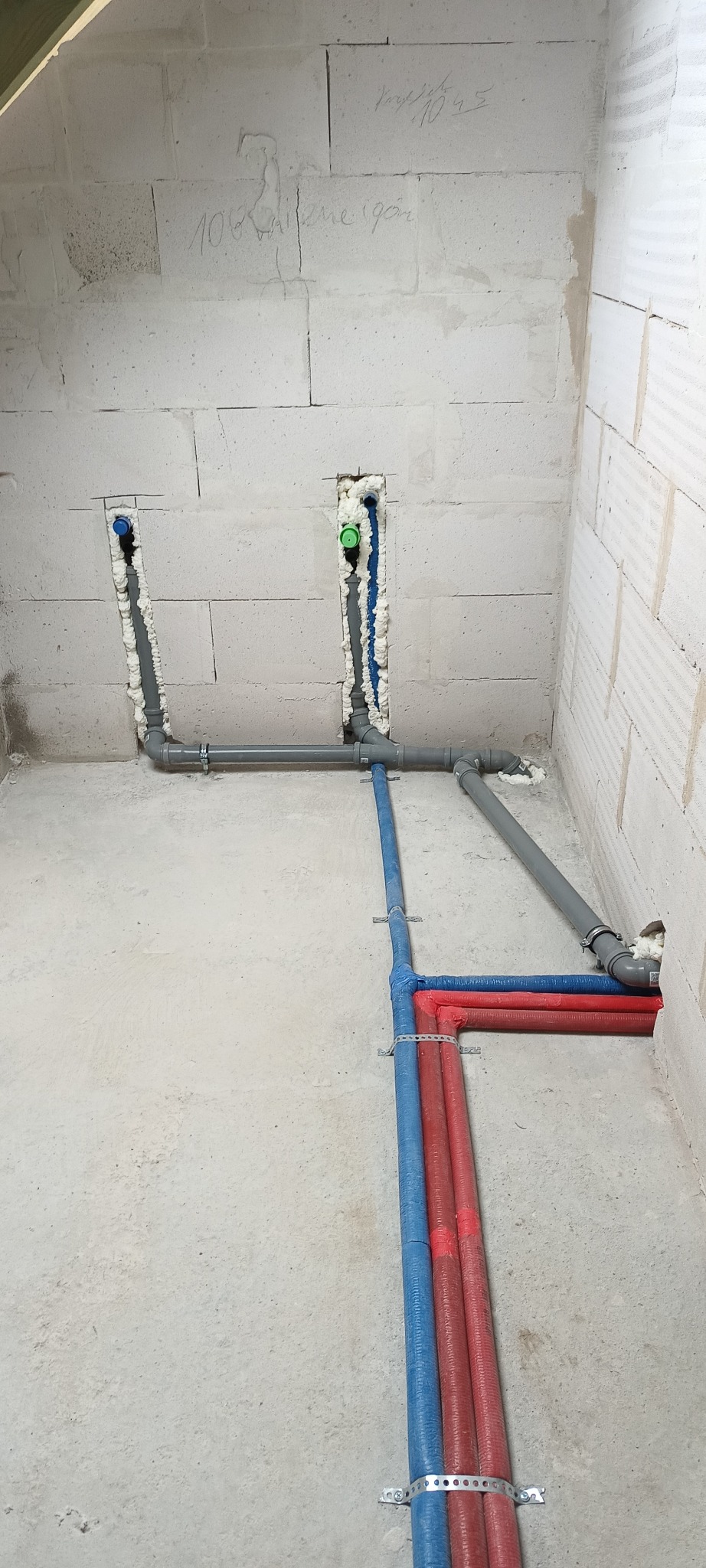 Surowa ściana z betonu komórkowego z wyciętymi bruzdami, w których umieszczono rury instalacji wodnej i grzewczej zabezpieczone pianką montażową, widoczne rury PEX w kolorze niebieskim i czerwonym...