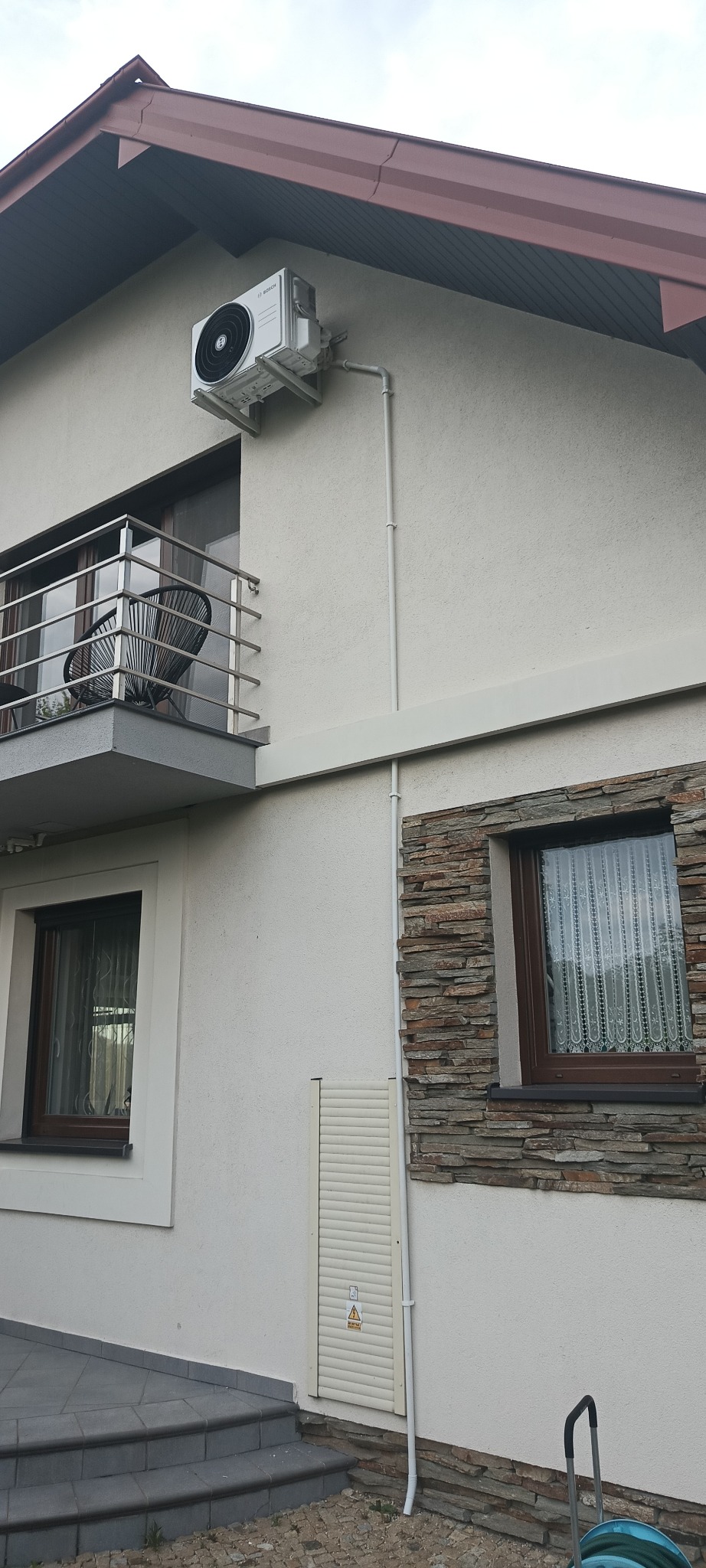 Zewnętrzna jednostka klimatyzacji zamontowana na ścianie domu z balkonem i oknem obłożonym kamieniem dekoracyjnym.