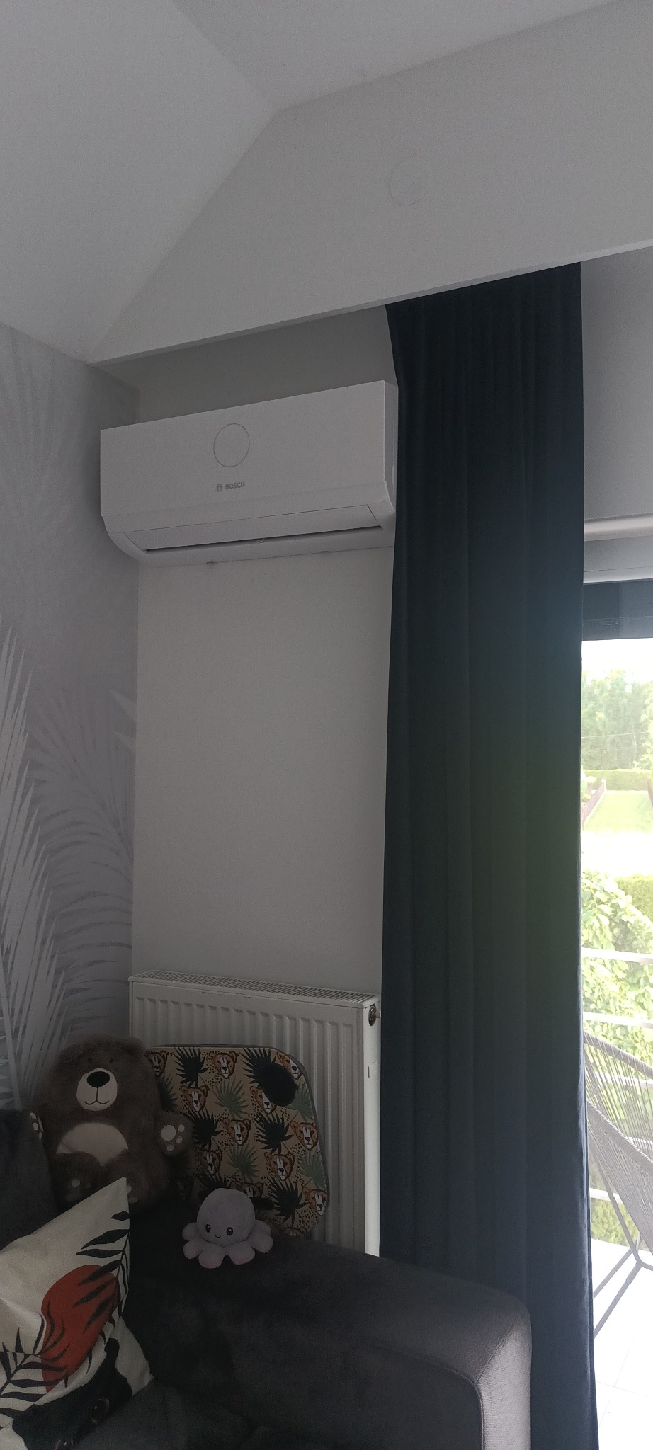 Biały klimatyzator Bosch zamontowany na ścianie w pokoju, widoczny grzejnik, zasłony i zabawki na kanapie.