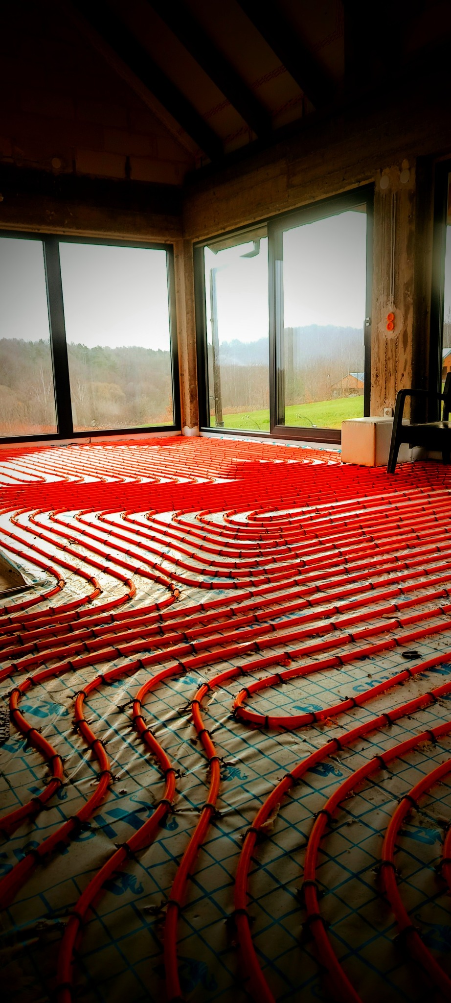 Pomarańczowe rury ogrzewania podłogowego ułożone spiralnie na izolacji z nadrukiem siatki w przestronnym, surowym wnętrzu z dużymi oknami i widokiem na zalesiony krajobraz.