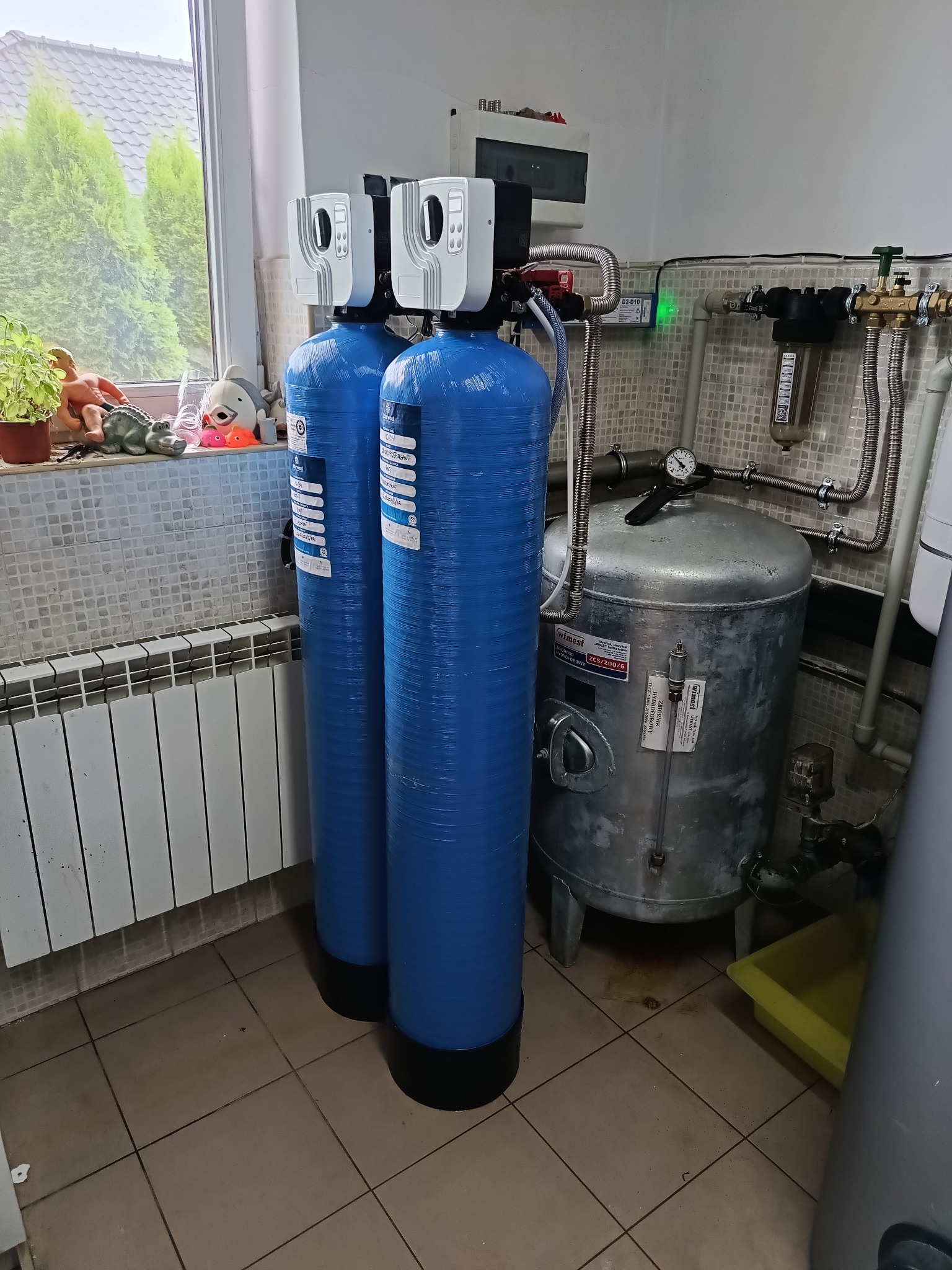 Dwa niebieskie filtry do wody z białymi głowicami, zbiornik hydroforowy marki Wimest, instalacja wodna, zabawki na parapecie okna w tle.