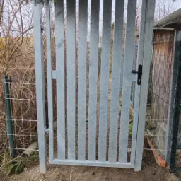 Szara, metalowa furtka z pionowych profili z czarną klamką w Wałbrzychu, widoczna spod gałęzi drzewa, prowadząca na teren działki.