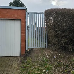 Prosta, ocynkowana furtka z pionowych profili, zamontowana między budynkiem garażu z cegły a gęstym krzewem, prowadząca na zarośnięty teren.