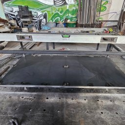 KLARIGA-WELD Maciej Burdziński - Metalowa rama z czarnym blatem na stole warsztatowym, obok szlifierka kątowa; w tle widoczny zniszczony samochód i graffiti na ścianie.